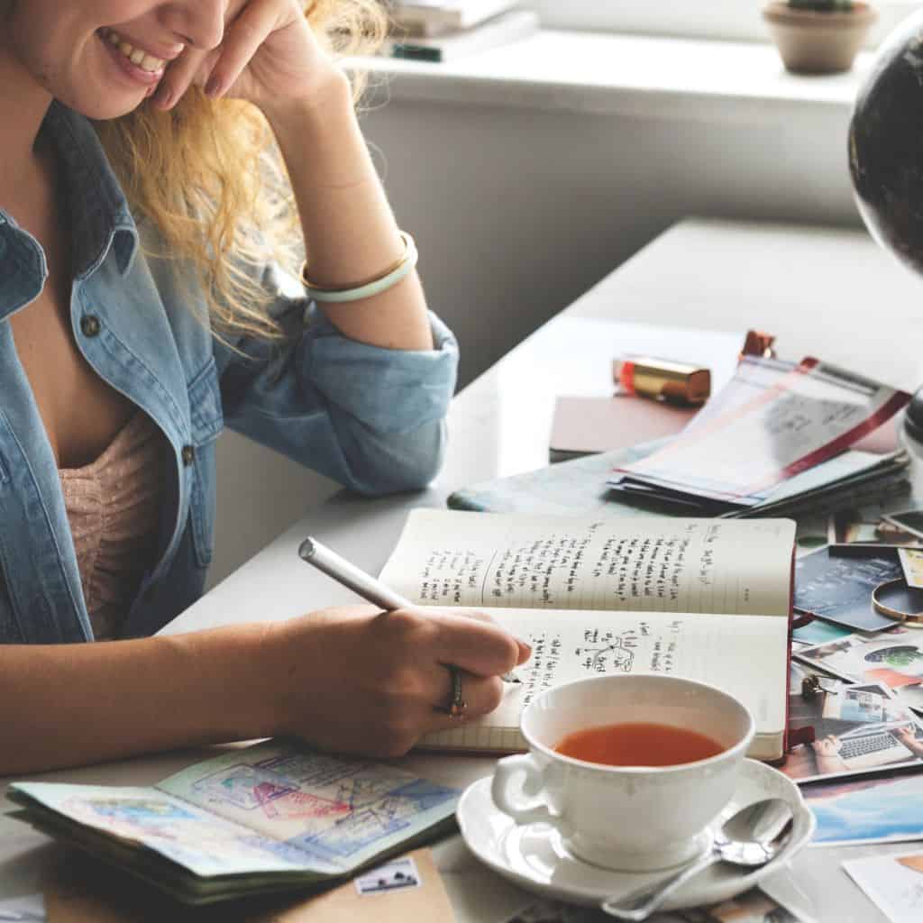 A woman sitting at a desk writing in her bucket list travel diary with a cup of tea beside her.
