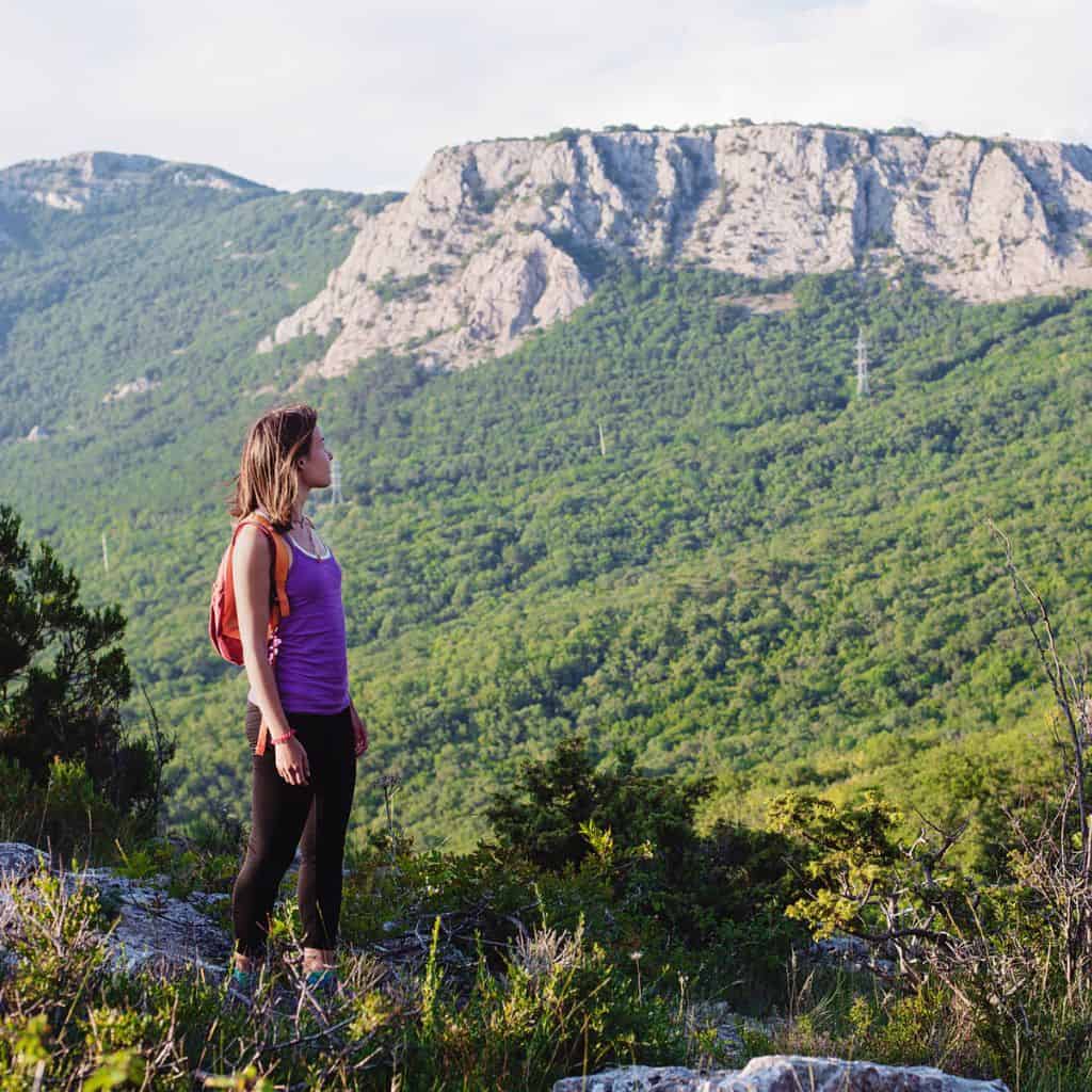 Young woman in a purple tank top hiking along a mountain side.
